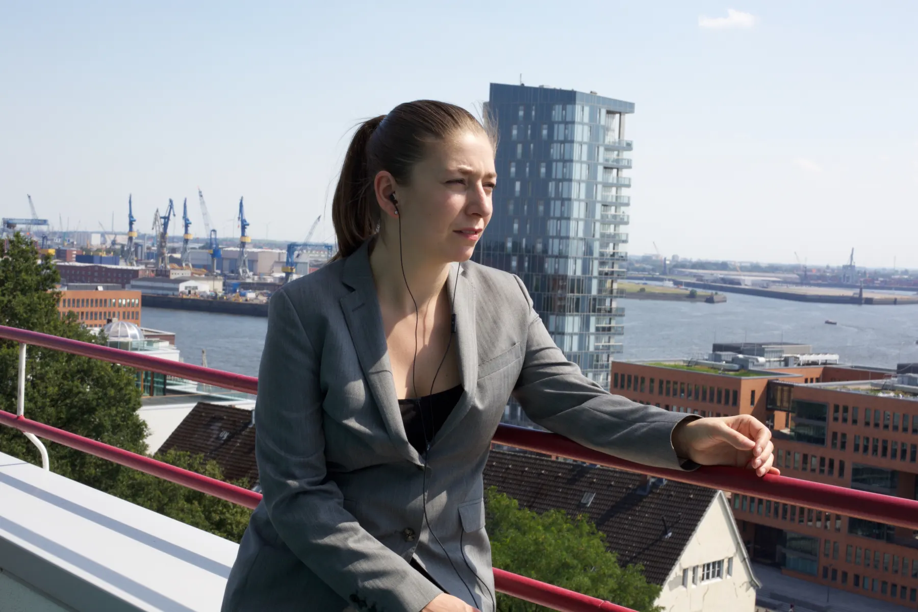 A woman in a grey blazer leans on a balcony railing above Hamburg harbour