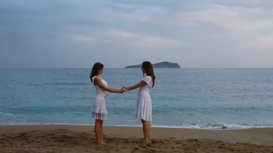 Two women in white dresses hold hands on a beach, an island on the horizon