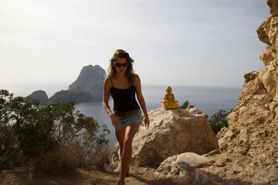 A woman climbs rocky cliffs, the islet Es Vedrà behind her
