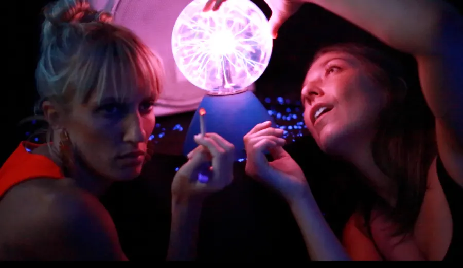 Two women gaze at a glowing plasma ball in violet light