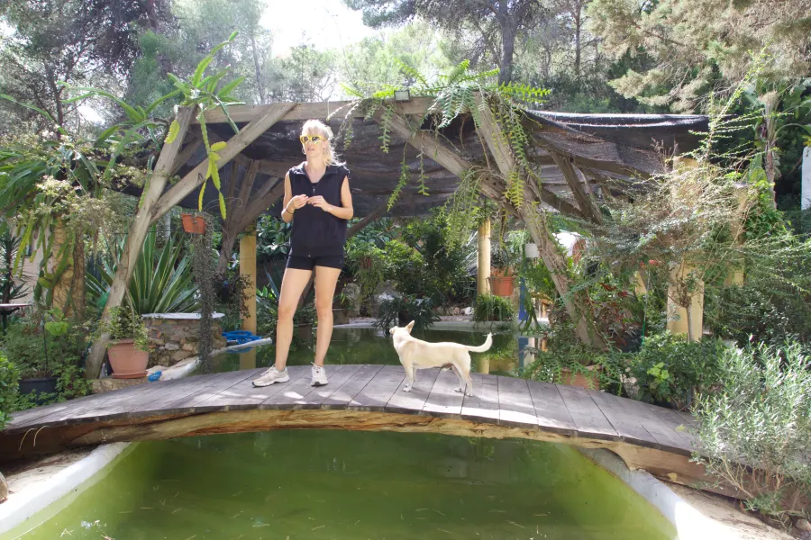 A woman stands on a wooden platform in a lush garden, a small dog beside her